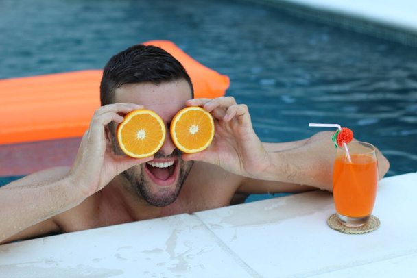 közeli kép egy jóképű fiatalemberről, aki fél narancsot tart a szeme előtt a medencében. - Fotó, kép