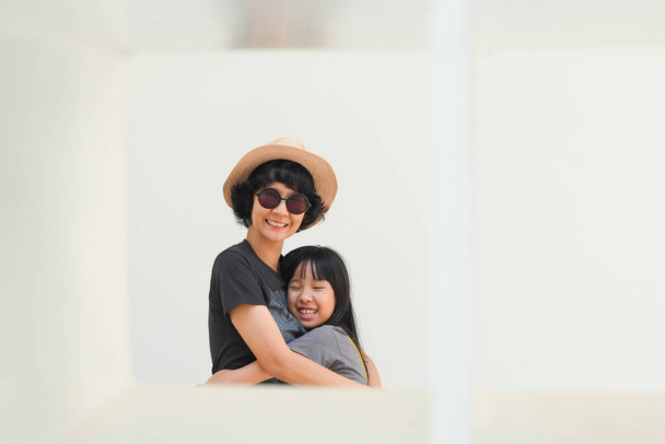 帽子と眼鏡をかけたTシャツを着たアジア系の母親は、白い背景の中でリラックスした幸せな気分で娘を抱きしめる. - 写真・画像