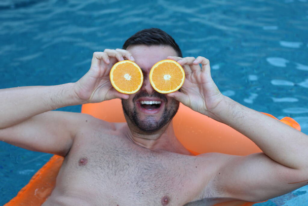close-up portrait of handsome young man floating on inflatable mattress holding halves of orange in front of eyes in swimming pool - Photo, Image