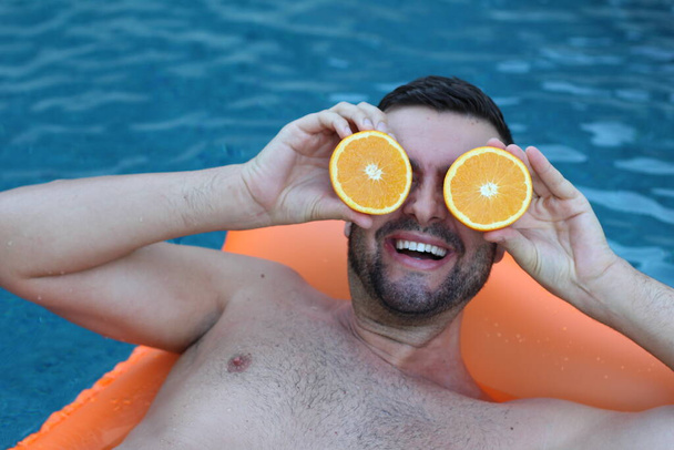 close-up portrait of handsome young man floating on inflatable mattress holding halves of orange in front of eyes in swimming pool - Фото, изображение
