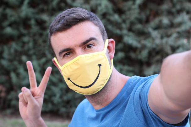 zbliżenie portret przystojny młody człowiek z medyczną maską dla covid ochrona z uśmiechem robienie selfie w zielonym parku, komedia koncepcja - Zdjęcie, obraz