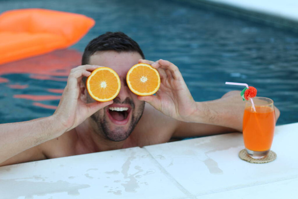 close-up portrait of handsome young man holding halves of orange in front of eyes in swimming pool - Fotoğraf, Görsel