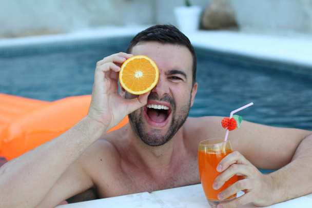 Nahaufnahme Porträt eines hübschen jungen Mannes, der im Schwimmbad die Hälfte der Orange vor dem Auge hält - Foto, Bild