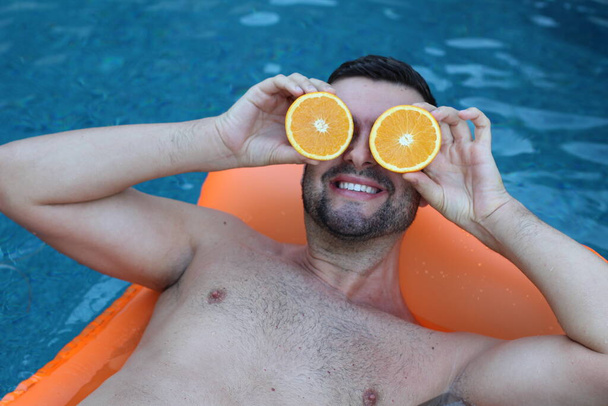close-up portrait of handsome young man floating on inflatable mattress holding halves of orange in front of eyes in swimming pool - Photo, Image
