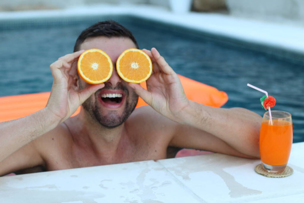 close-up portrait of handsome young man holding halves of orange in front of eyes in swimming pool - Photo, Image