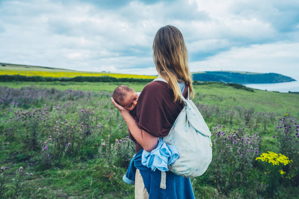 A young mother is standing by the seaside in a meadow with her baby in a sling - Foto, Imagen