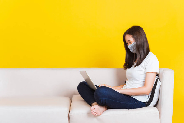 Portrait Asian of beautiful young woman sitting on sofa wearing medical face mask protective she work from home with laptop computer during Coronavirus studio shot isolated on yellow background - Foto, imagen