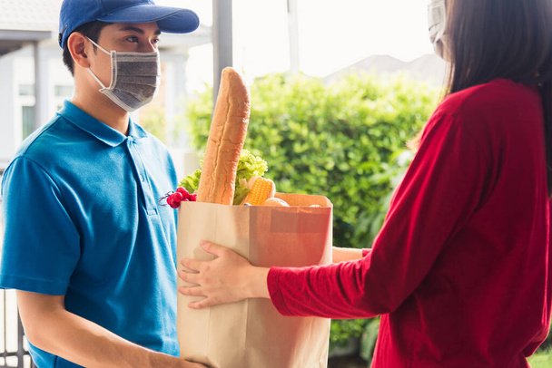 Asian young delivery man in uniform wear protective face mask he making grocery service giving fresh food to woman customer receiving front house under pandemic coronavirus, Back to new normal concept - Фото, изображение