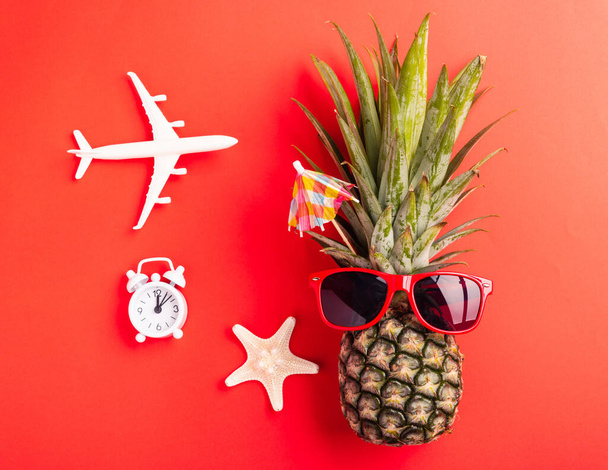 Summer Pineapple Day Concept; Top view flat lay of funny pineapple wear red sunglasses; model plane; starfish and clock alarm isolated on red background; Holiday summertime in tropical - Фото, зображення