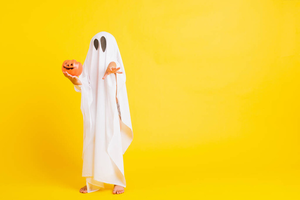 Funny Halloween Kid Concept, little cute child with white dressed costume halloween ghost scary he holding orange pumpkin ghost on hand, studio shot yellow on white background - Photo, Image