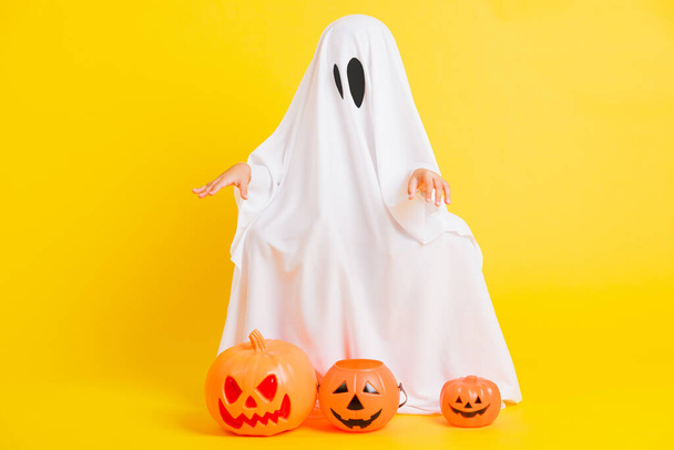 Funny Halloween Kid Concept, full body of a little cute child with white dressed costume halloween ghost scary he holding orange pumpkin ghost, studio shot yellow on white background - Foto, Imagem