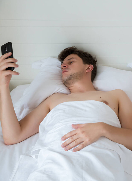 Man in bed with cellphone watching latest new. Young man lying in white sheets bed in the hotel and watching social media network by phone. - Фото, изображение