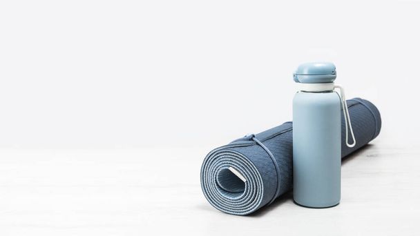 Rolled blue yoga mat and blue water bottle on grey wooden surface. Gender neutral fitness yoga and exercise concept with copy space. Active lifestyle. Workout at home or gym banner - Φωτογραφία, εικόνα