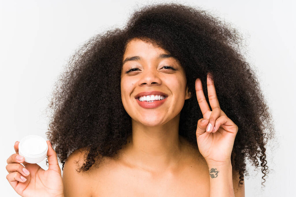 Jeune femme afro tenant une crème hydratante isolée montrant signe de victoire et souriant largement. - Photo, image