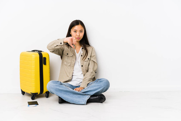 Joven viajera china sentada sosteniendo una tarjeta de embarque mostrando un gesto de aversión, con los pulgares hacia abajo. Concepto de desacuerdo. - Foto, Imagen