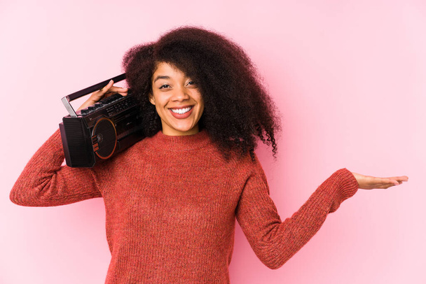 Joven afro mujer sosteniendo un cassete aislado mostrando un espacio de copia en una palma y sosteniendo otra mano en la cintura. - Foto, Imagen