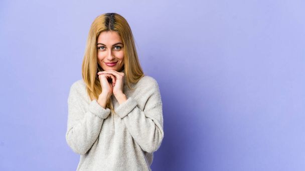 Young blonde woman isolated on purple background keeps hands under chin, is looking happily aside. - Photo, Image