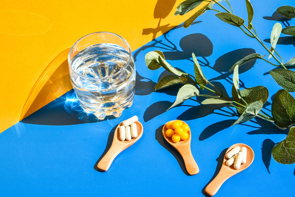 Bicchiere d'acqua e pillole su cucchiaio di legno su sfondo blu e giallo. Luce e ombre dure. Moderno concetto di minimalismo creativo isometrico. Vitamine e prebiotici, probiotici. Dose di vitamina autunnale. Eucalipto - Foto, immagini