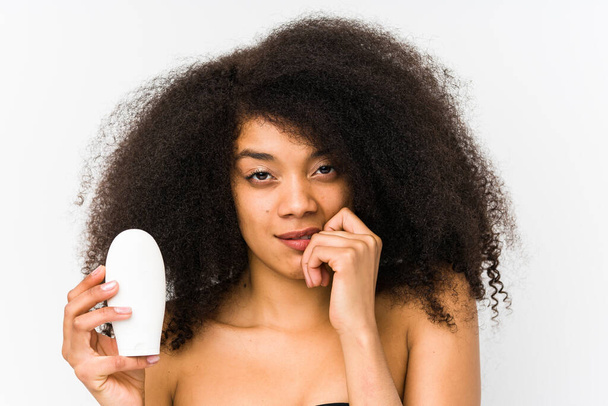 Mujer afro joven sosteniendo una crema hidratante aislada mordiendo las uñas, nerviosa y muy ansiosa. - Foto, imagen