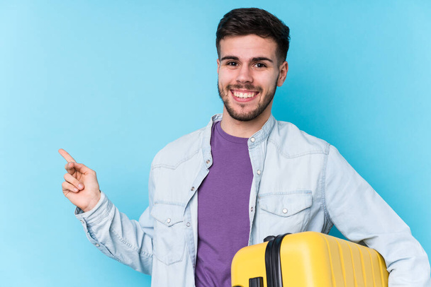 Young caucasian traveler man holding a suitcase isolated smiling and pointing aside, showing something at blank space. - Foto, Imagem