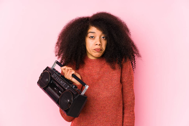 Joven afro mujer sosteniendo un cassete aislado se encoge de hombros y los ojos abiertos confundido. - Foto, imagen