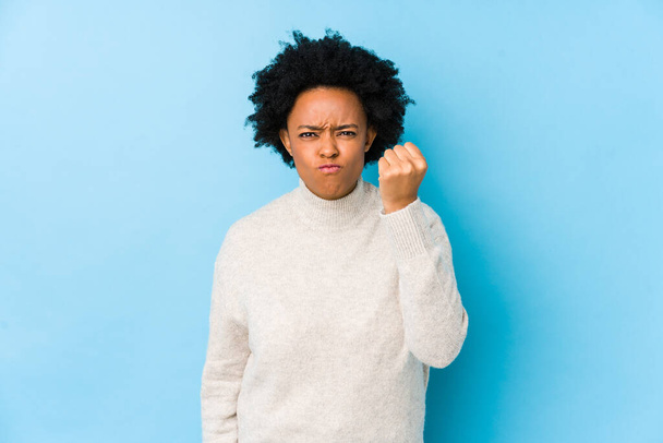 Femme afro-américaine d'âge moyen sur un fond bleu isolé montrant poing à caméra, expression faciale agressive. - Photo, image