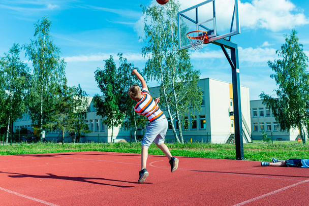 Aantrekkelijke blanke kleine basketbalspeler springt om de bal in de basket hoepel te gooien op een buiten rode baan - Foto, afbeelding