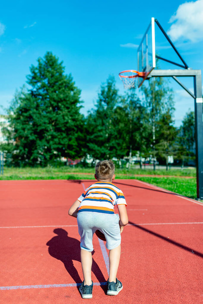 Houkutteleva valkoihoinen pikku koripalloilija koulutusta ulkona punainen kenttä takaisin näkymä poika hyppää heittää pallon koriin - Valokuva, kuva