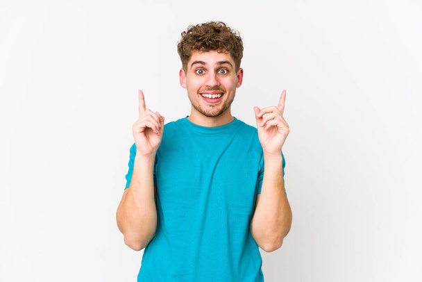 Jovem loiro cabelo encaracolado caucasiano homem isolado indica com ambos os dedos dianteiros para cima mostrando um espaço em branco. - Foto, Imagem
