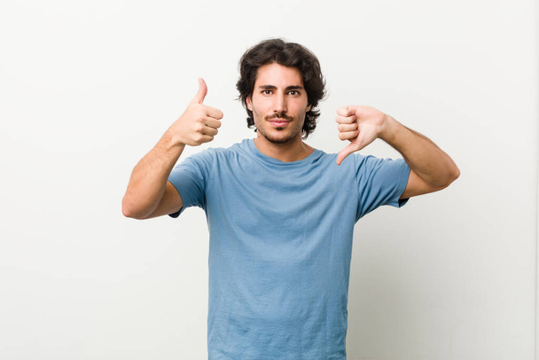 Young handsome man against a white background showing thumbs up and thumbs down, difficult choose concept - Photo, Image