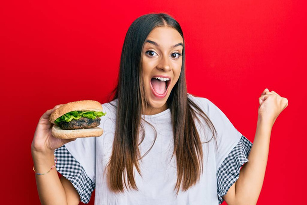 Junge hispanische Frau isst stolz Hamburger und feiert Sieg und Erfolg mit erhobenen Armen  - Foto, Bild