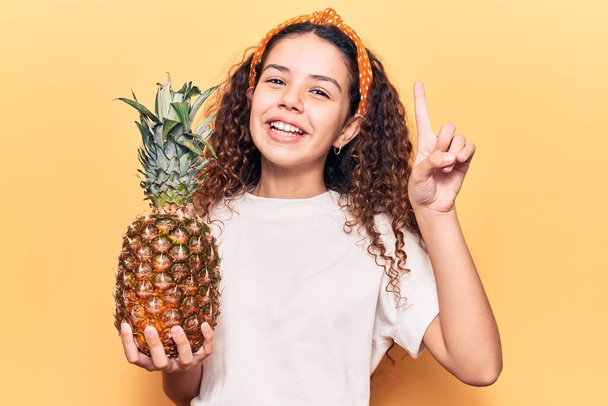 Красива дівчинка з кучерявим волоссям тримає ананас, посміхаючись з ідеєю або питанням, спрямованим пальцем з щасливим обличчям, номер один
  - Фото, зображення