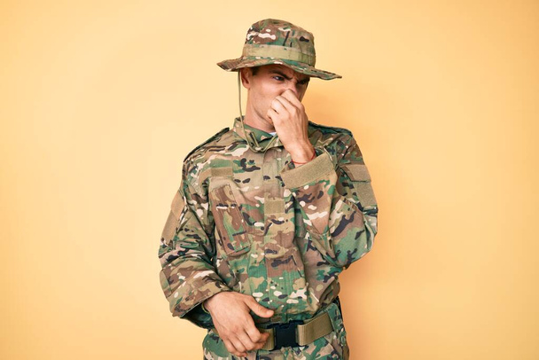 Молодой красивый мужчина в камуфляжной армейской форме, пахнущий чем-то вонючим и отвратительным, невыносимым запахом, затаив дыхание с пальцами на носу. плохой запах  - Фото, изображение