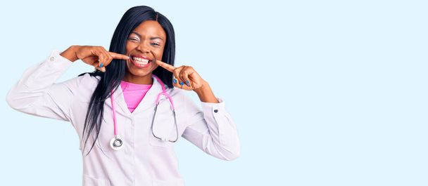 Doktor steteskop takan, gülümseyen, parmaklarıyla ve ağzıyla işaret eden genç Afro-Amerikalı bir kadın. diş sağlığı kavramı.  - Fotoğraf, Görsel
