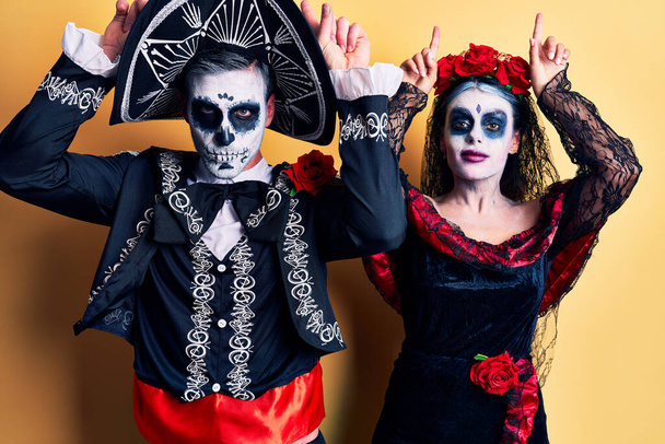 Молодая пара, одетая в мексиканский день мертвого костюма над желтым, делает смешной жест с пальцем над головой, как рога быка.  - Фото, изображение