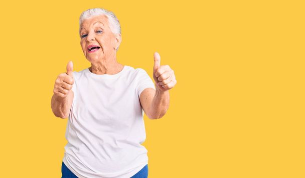 Senior beautiful woman with blue eyes and grey hair wearing casual white tshirt approving doing positive gesture with hand, thumbs up smiling and happy for success. winner gesture.  - Foto, Bild