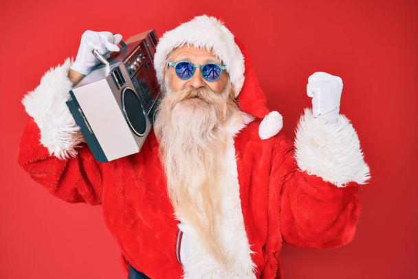 Old senior man wearing santa claus costume and boombox screaming proud, celebrating victory and success very excited with raised arms  - Photo, Image