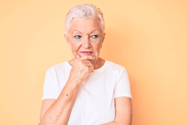 Senior beautiful woman with blue eyes and grey hair wearing classic white tshirt over yellow background with hand on chin thinking about question, pensive expression. smiling with thoughtful face. doubt concept.  - Photo, Image