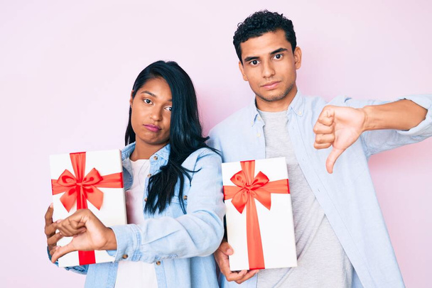 Schönes lateinisches junges Paar hält Geschenk für Jubiläumsfeier mit wütendem Gesicht, negatives Zeichen zeigt Abneigung mit gesenktem Daumen, Ablehnungskonzept  - Foto, Bild