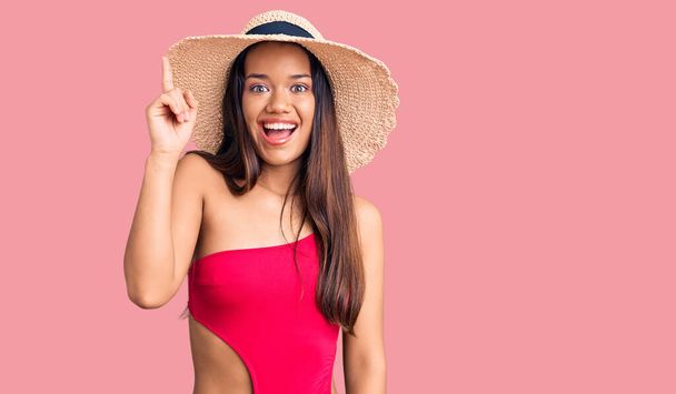 Fiatal, gyönyörű latin lány fürdőruhát és nyári kalapot visel, ujjal mutogatva a sikeres ötletre. kilépett és boldog volt. Egyes számú..  - Fotó, kép