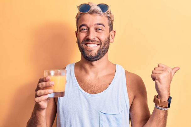 Young handsome blond man with beard on vacation drinking glass of healthy orange juice pointing thumb up to the side smiling happy with open mouth - Foto, Imagen