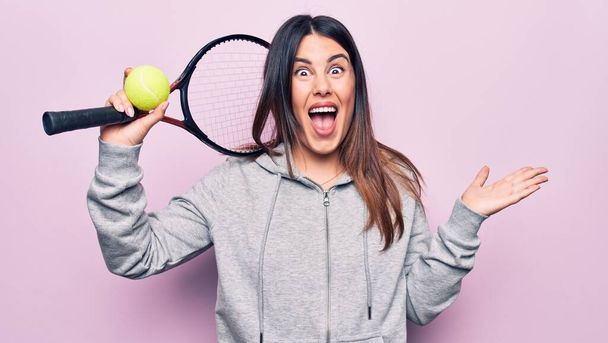 Jonge mooie sportvrouw spelen tennis met behulp van racket en bal over roze achtergrond vieren prestatie met een vrolijke glimlach en winnaar uitdrukking met opgeheven hand - Foto, afbeelding