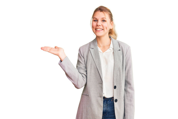 Jovem mulher loira bonita vestindo jaqueta elegante sorrindo alegre apresentando e apontando com palma da mão olhando para a câmera.  - Foto, Imagem