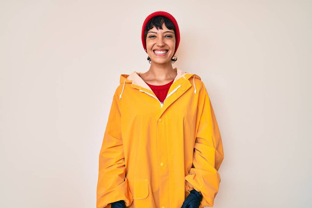 Beautiful brunettte woman wearing yellow raincoat looking positive and happy standing and smiling with a confident smile showing teeth  - Fotoğraf, Görsel