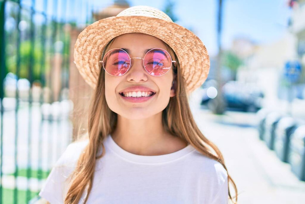 Молодая красивая блондинка-кавказка улыбается на открытом воздухе в солнечный день в летней шляпе и розовых солнечных очках - Фото, изображение