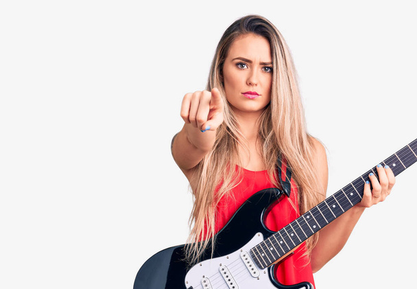 Fiatal gyönyörű szőke nő játszik elektromos gitár mutató ujjal a kamera és az Ön számára, magabiztos gesztus néz komoly  - Fotó, kép