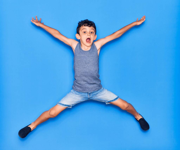 Αξιολάτρευτο παιδί που φοράει καθημερινά ρούχα. Άλμα με τα χέρια ανοιχτά πάνω από απομονωμένο μπλε φόντο - Φωτογραφία, εικόνα
