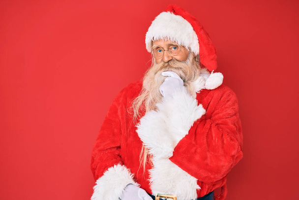 Oude senior man met grijs haar en lange baard draagt traditionele kerstman kostuum kijken zelfverzekerd naar de camera met glimlach met gekruiste armen en hand opgeheven op kin. positief denken.  - Foto, afbeelding