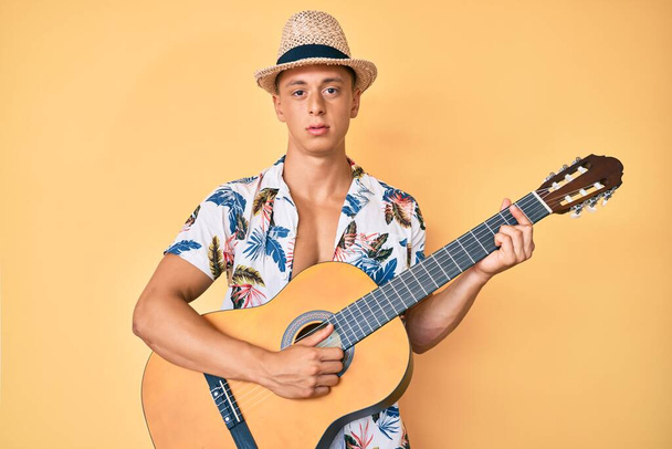 Νεαρό ισπανόφωνο αγόρι που φοράει καλοκαιρινό στυλ παίζοντας κλασική κιθάρα χαλαρό με σοβαρή έκφραση στο πρόσωπο. απλή και φυσική κοιτάζοντας την κάμερα.  - Φωτογραφία, εικόνα