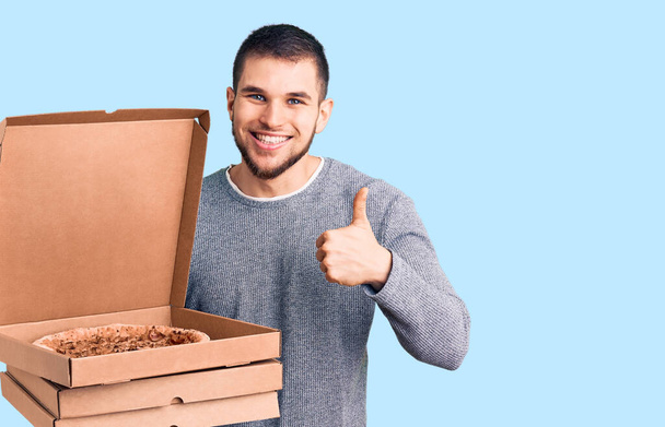 Junger gutaussehender Mann mit Lieferung italienischer Pizzakartons, der glücklich und positiv lächelt, Daumen hoch und Zeichen der Anerkennung  - Foto, Bild